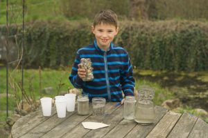 Ein Junge steht im Garten hinter einem Tisch. Darauf stehen vier Plastikbecher, zwei Gläser mit jeweils grobem und feinem Sand.