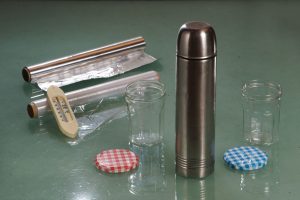 Alufolie, Plastikfolie, Thermometer, zwei Marmeladengläser mit Deckeln, und eine Thermoskanne mit warmem Wasser stehen auf einem Tisch.