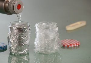 Eine Thermoskanne gibt Wasser in ein Marmeladenglas in Alu-Folie. Ein anderes mit Plastikfolie umwickeltes Glas steht daneben.