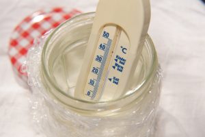 Ein Thermometer steht in einem mit Wasser gefülltem und mit Folie umwickelten Marmeladenglas.