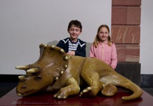 Zwei Kinder sitzen hinter dem Modell eines Triceratops-Baby.