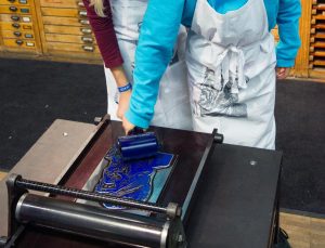 Zwei Kinder bestreichen eine Druckform mit blauer Farbe.  