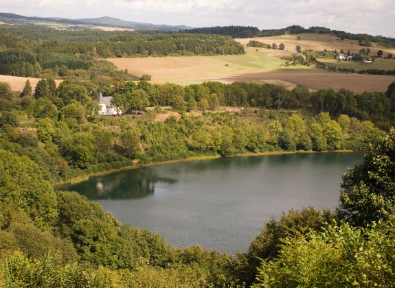Ein Landschaft mit See in der Eifel. Am Ufer steht ein kleines Haus.
