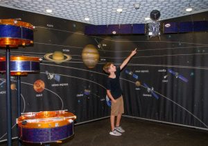 Ein Junge zeigt auf ein Modell des Satelliten Rosetta. Er steht in einem Raum mit Modellen vom Sonnensystem.