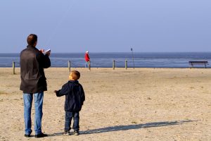 Ein Mann lässt am Strand einen Drachen steigen. Neben ihm steht ein kleiner Junge. 