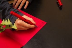 Ein Junge schreibt mit Füller eine Weihnachtskarte auf ein rotes Papier.
