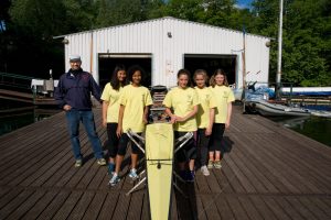 Fünf Kinder aus einem Ruderclub stehen auf dem Steg mit ihrem Boot. Daneben steht ihr Trainer Dirk Schreiber.