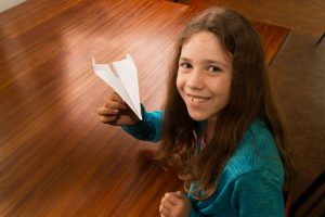 Ein junges Mädchen hält einen weißen Papierflieger hoch. Sie sitzt an einem braunen Tisch.