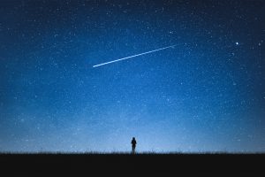 Ein blauer Nachthimmel mit Sternen und einer Sternschnuppe. Ein kleiner Mensch beobachtet den Himmel.