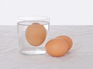 Ein Ei schwimmt in einem Glas Wasser. Daneben liegen zwei andere Eier: Ein Test, ob das Ei noch frisch ist.