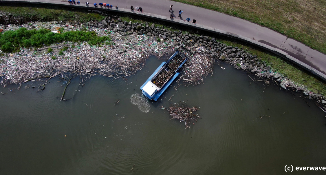 Eine asphaltierte und mit Steinen bewehrte Uferstraße ist von oben fotografiert. Am Ufer hat sich eine große Menge Treibgut, Müll und Plastik angesammelt. Das Everwave Boot räumt diesen Müll auf.