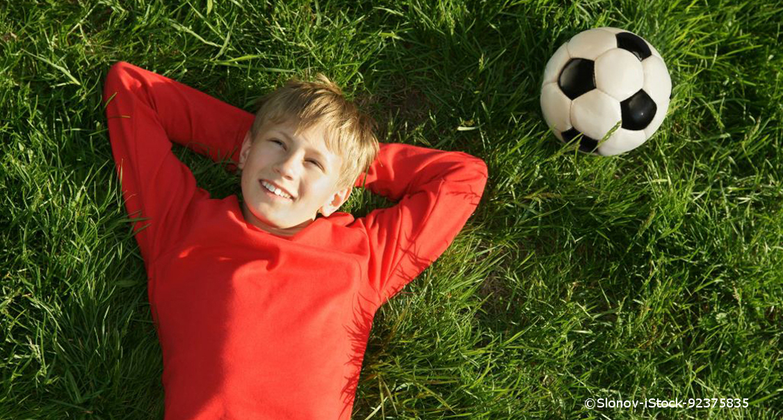 Ein blonder Junge in einem roten Fußballtrikot liegt auf einem Rasen.