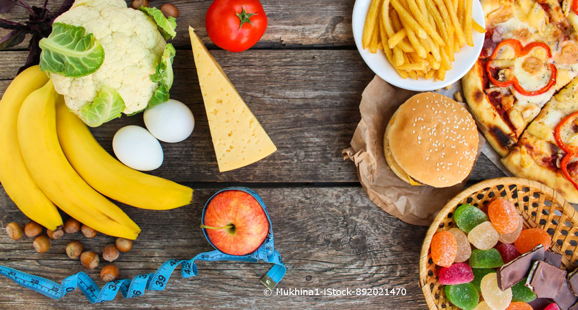 Ein Tisch mit slow food und fast food: Hamburger, Bananen, Süßigkeiten, ein Käse, Eier, Blumenkohl und Pizza.