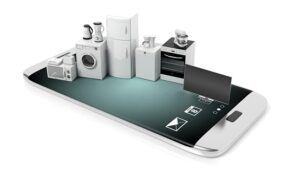 Viele Elektrogeräte stehen auf einem großen Smartphone. Unter den Geräten lassen sich eine Waschmaschine, ein Kühlschrank oder ein Fernseher finden.