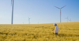 Ein Mann mit einem weißen Bauhelm steht in einem Weizenfeld und macht sich Notizen. Im Hintergrund sind Windräder zu erkennen, die in der Landschaft stehen. Der Hintergrund ist ein wolkenfreiere blauer Himmel.