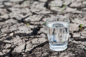 Gegen Wassermangel: ein Glas klares Wasser auf trockenem Boden.